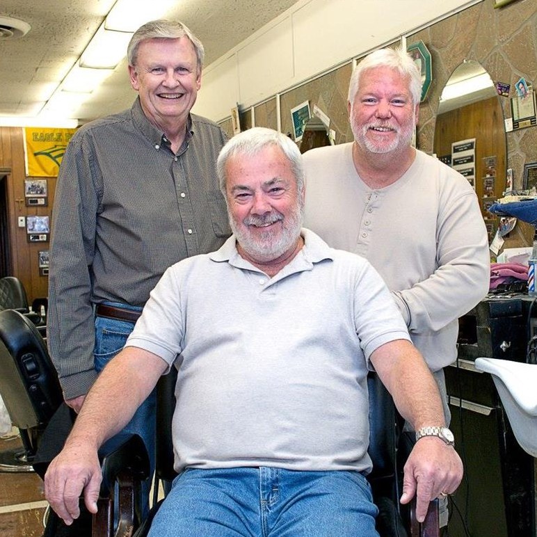 The Busy Bee’s three barbers in the shop, one seated in a barber chair and two standing behind him