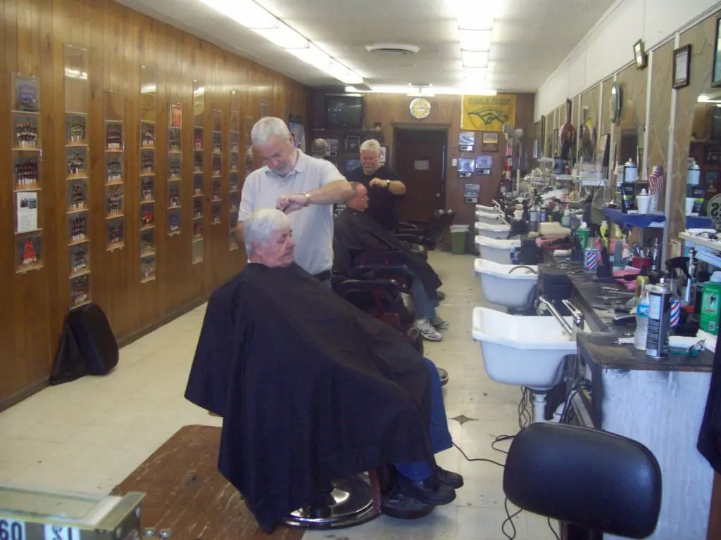 Two barbers at work in the long, narrow shop, with team photos along the paneled wall